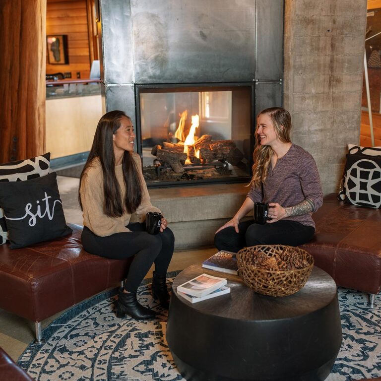 Two Gravity Haus Truckee-Tahoe guests enjoy hot drinks and chat by the fire in the lobby.
