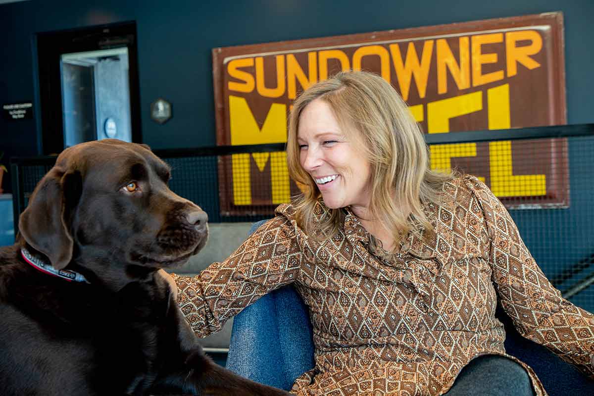Kerry Crandell relaxes and pets her chocolate lab at Garvity Haus Winter Park with historic Sundowner Motel sign in background.